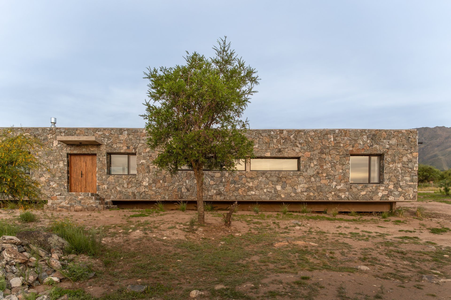 Casa revestida de piedra con un pequeño árbol al frente, situada en un paisaje seco y desértico.