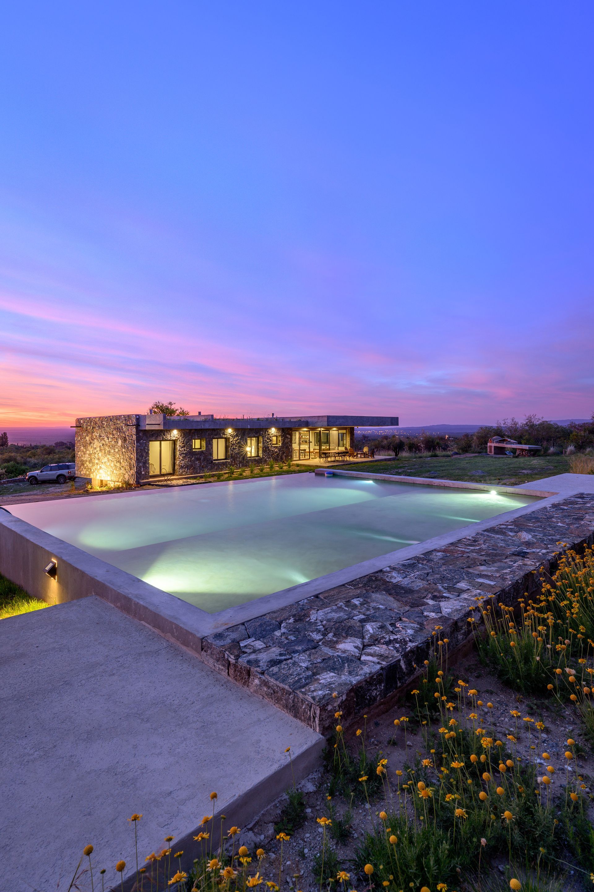 Piscina infinita al anochecer, con una casa iluminada al fondo.