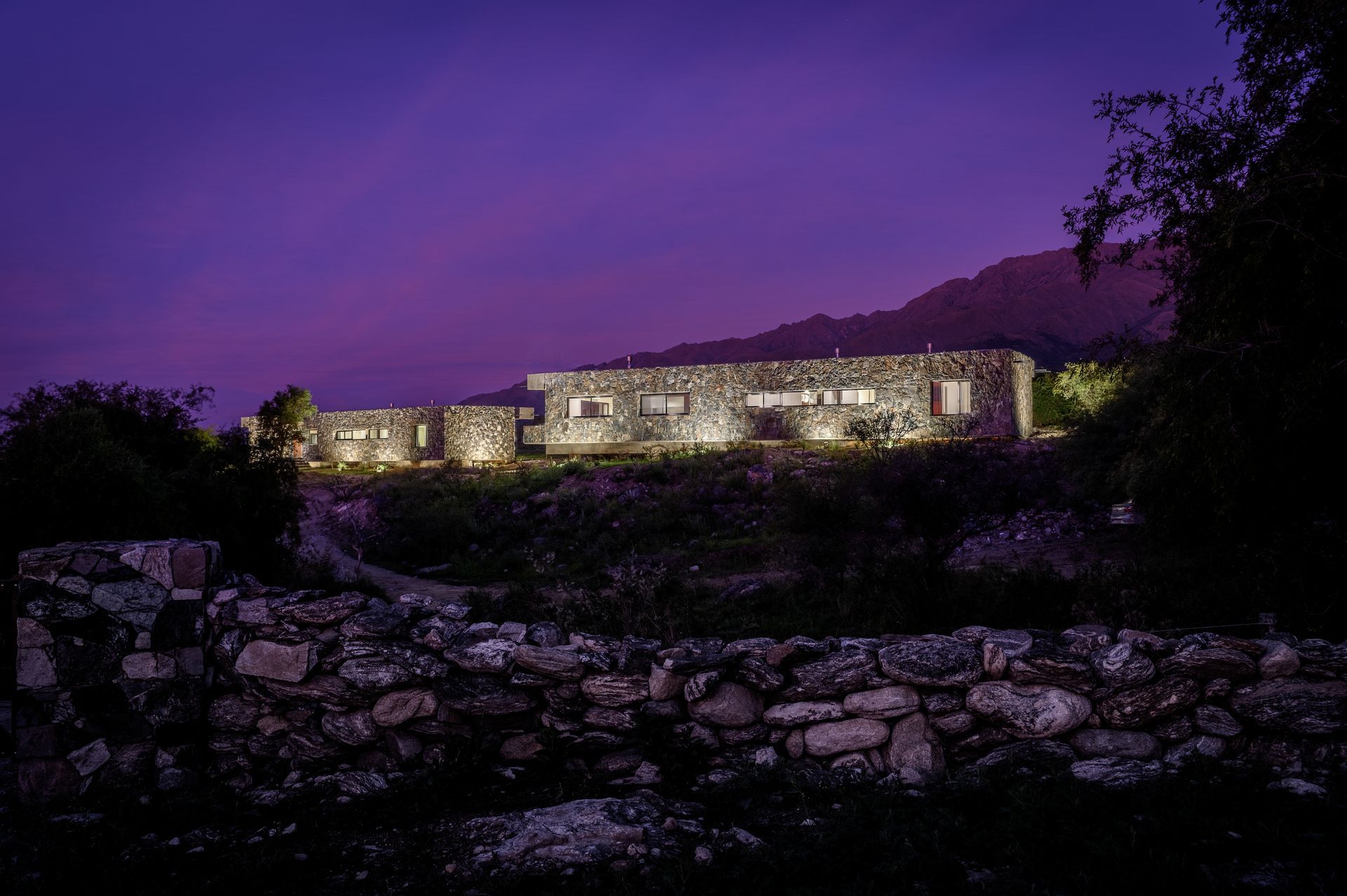 Casa de piedra al anochecer, iluminada desde dentro, contra un cielo púrpura y una colina.