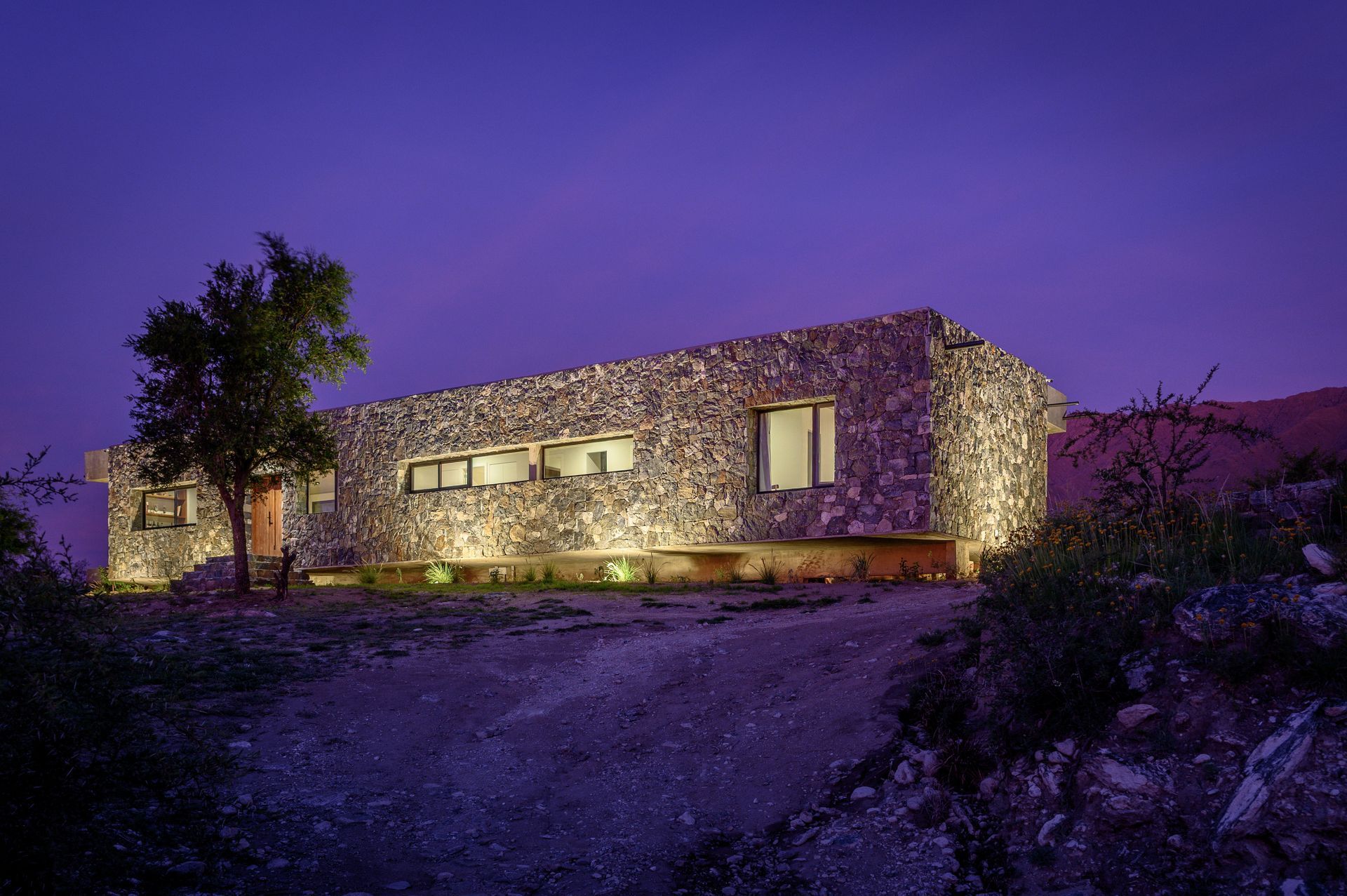Casa de piedra iluminada de noche, en un entorno natural, con un camino de acceso. Cielo morado.