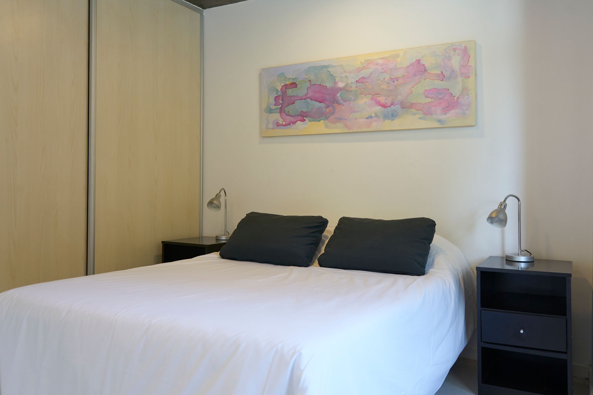 Dormitorio con ropa de cama blanca, almohadas y mesitas de noche negras. Una obra de arte cuelga sobre la cama.