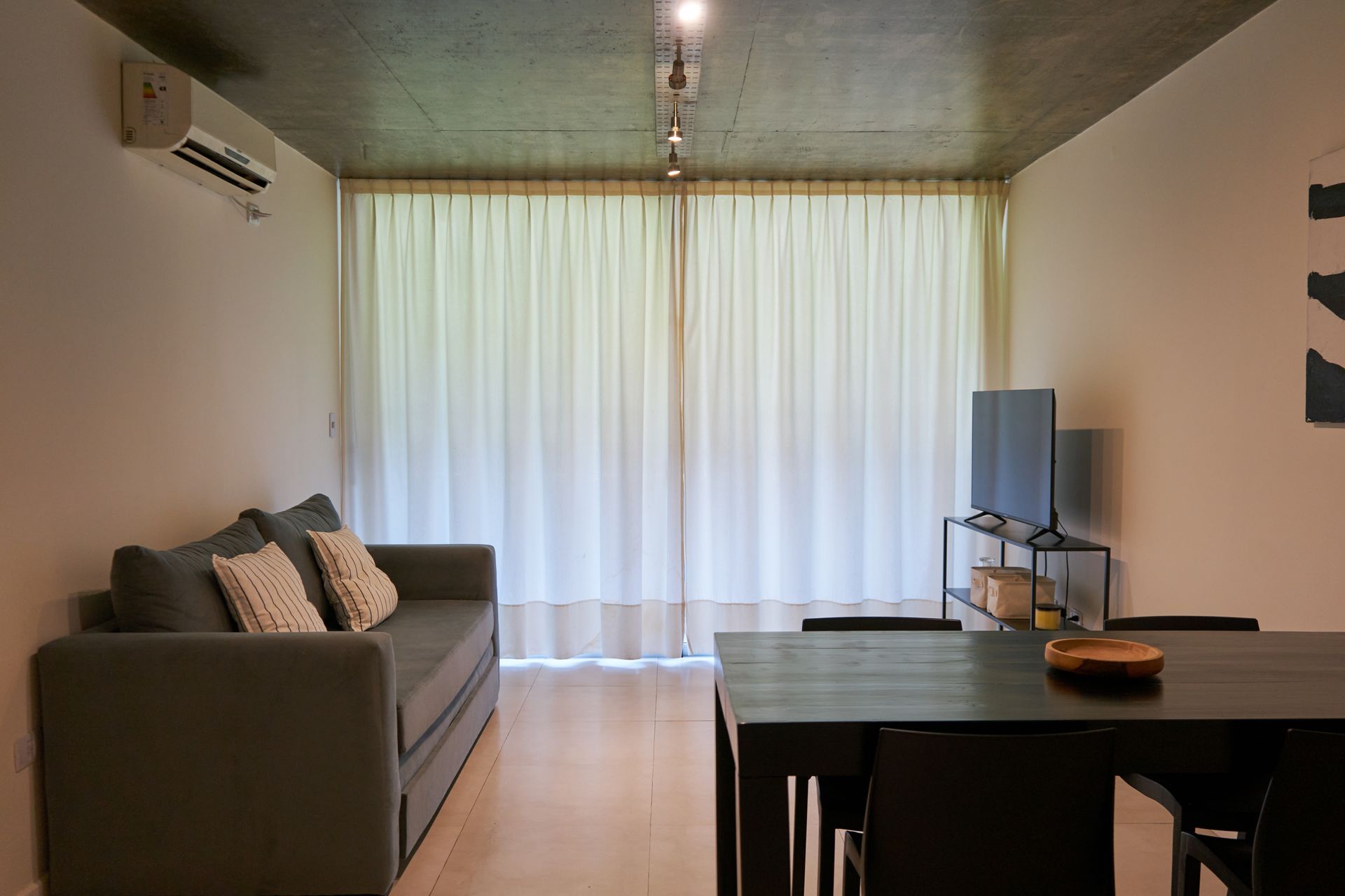 Sala de estar con sofá gris, mesa de comedor y TV frente a un gran ventanal con cortinas transparentes.