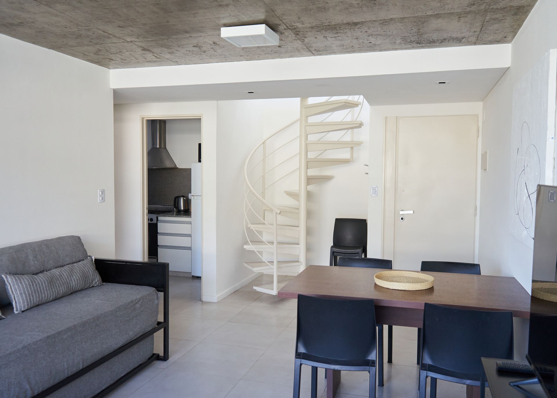 Apartamento moderno con escalera de caracol, sala de estar y comedor. Sofá gris, sillas negras y cocina a la vista.