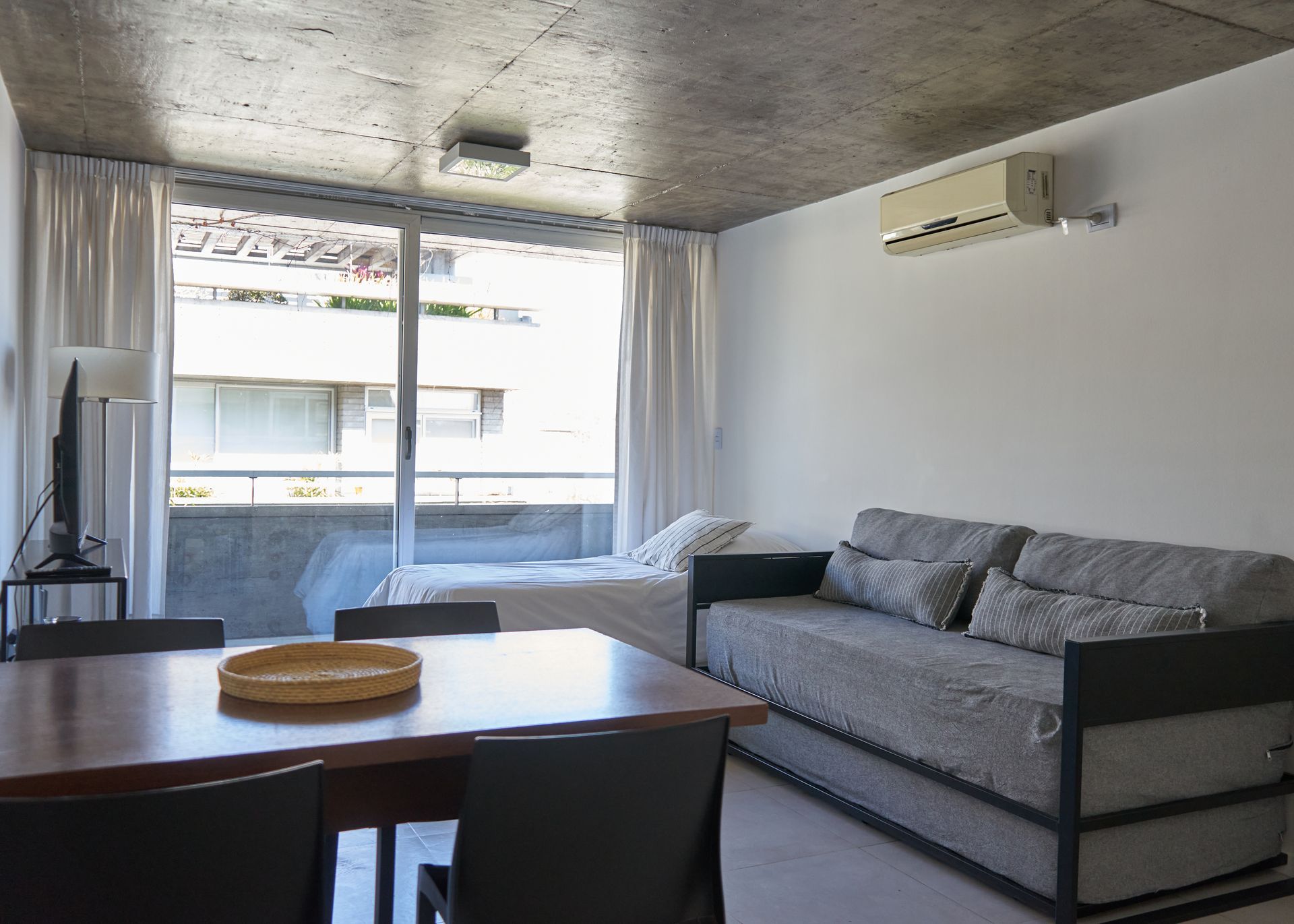 Pequeño apartamento con zona de comedor, cama y sofá cerca de una gran ventana.
