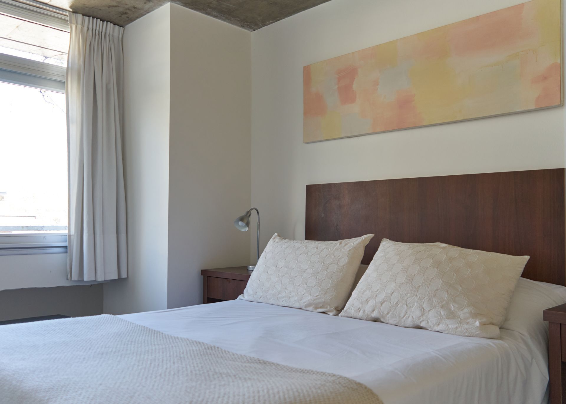 Dormitorio con cama, almohadas, obras de arte, ventana y mesita de noche.