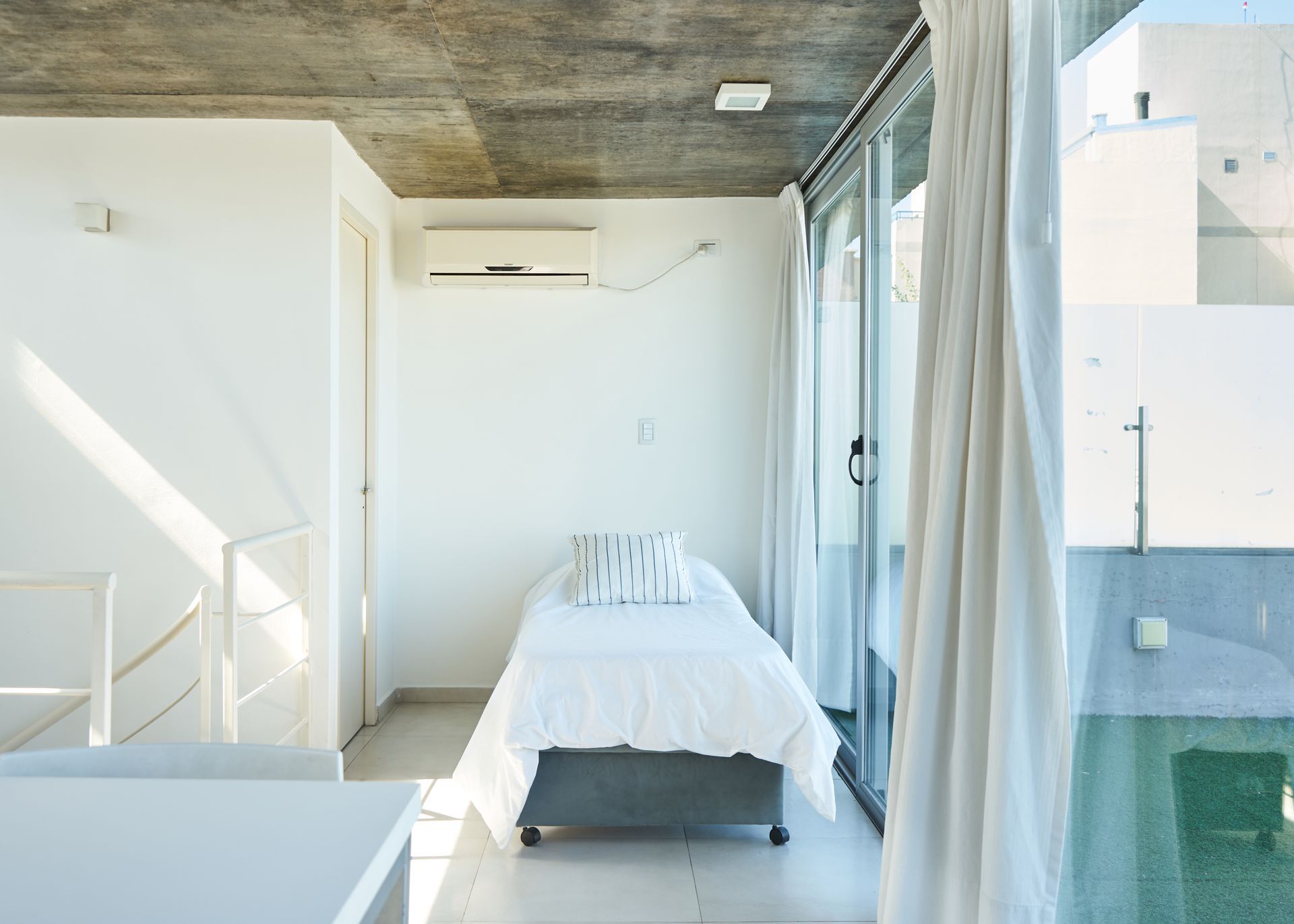 Dormitorio con cama individual, paredes blancas, techo de hormigón y gran ventanal.
