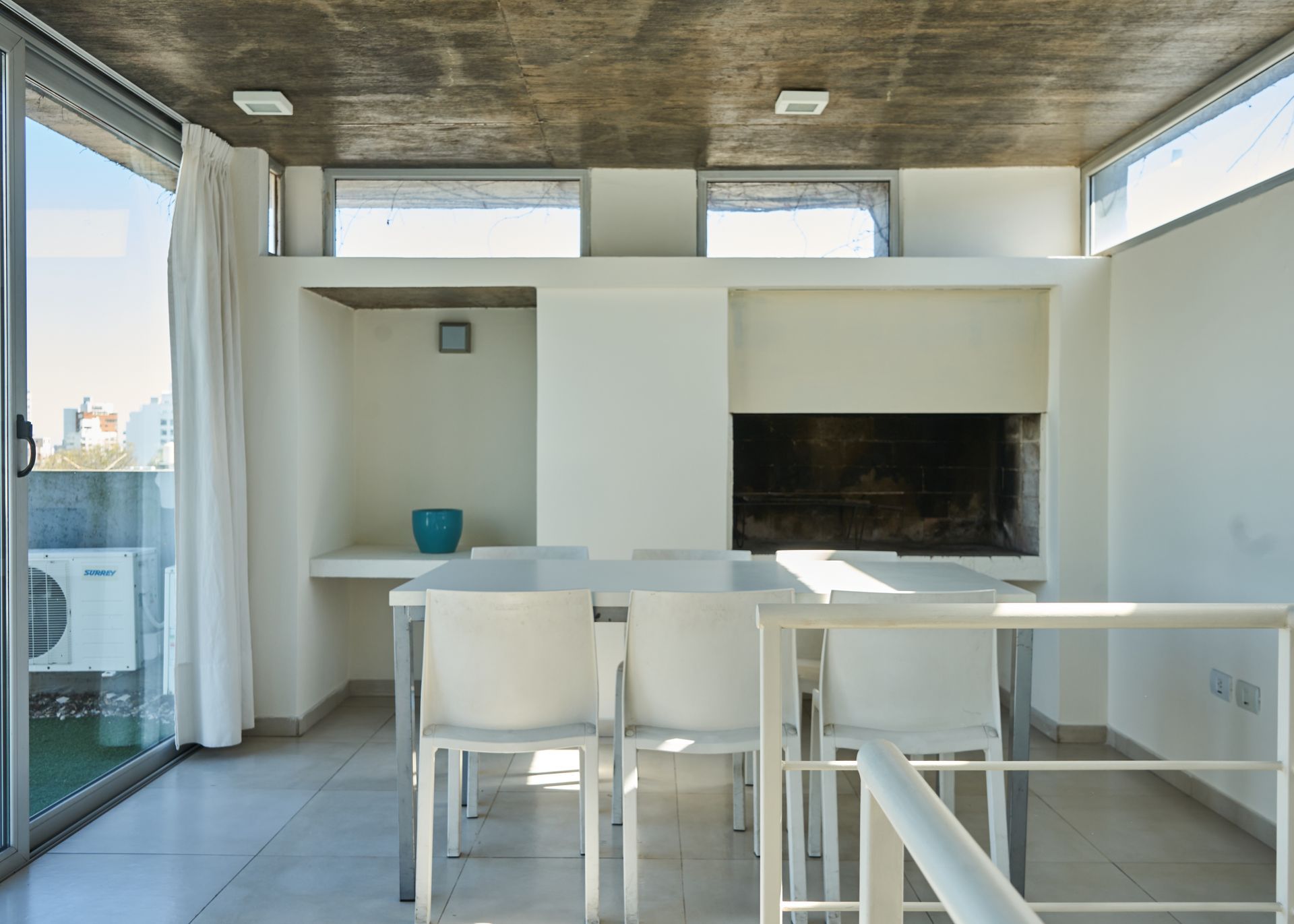 Comedor en azotea con muebles blancos, parrilla incorporada, techo de concreto y puerta corrediza de vidrio.