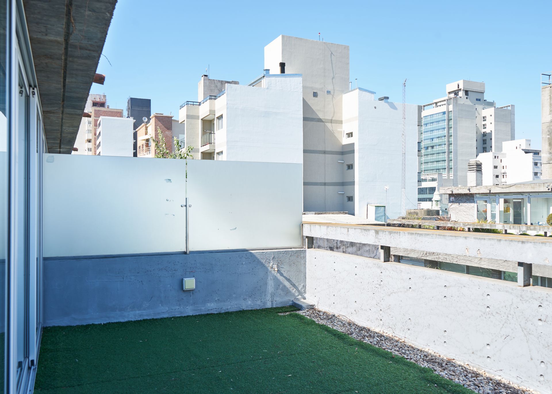Patio en azotea con césped artificial y edificios al fondo bajo un cielo despejado.