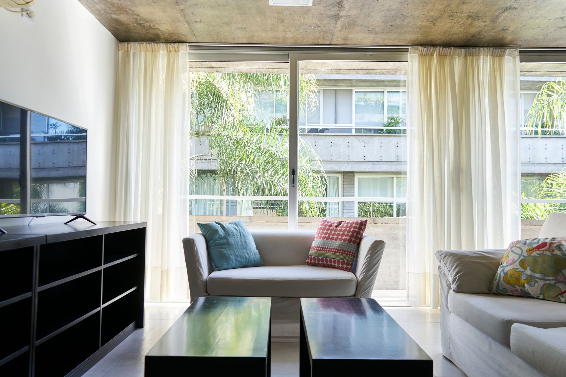 Sala de estar con sofá blanco, almohadas azules y estampadas, mesas de café negras y ventana alta con cortinas transparentes.