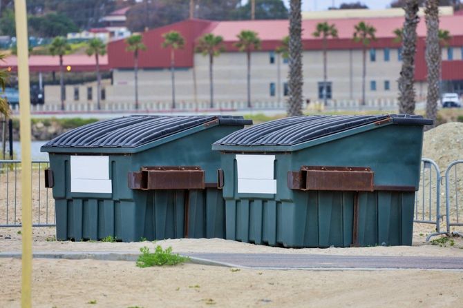 Two green dumpsters with black lids and white signs beside a sandy lot and building.