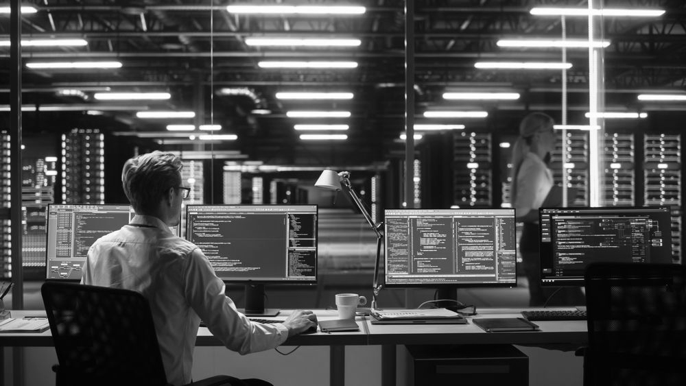 Man at computer in server room; woman walks past servers. Blue light beams.