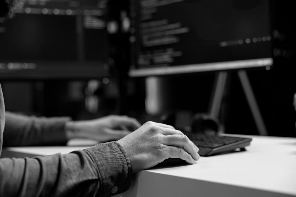 A close-up of a person's hands typing on a keyboard at a desk, with computer screens displaying code in the background.