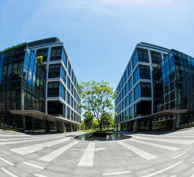 Modern glass office buildings framing a bright courtyard with a tree and crosswalk under a blue sky