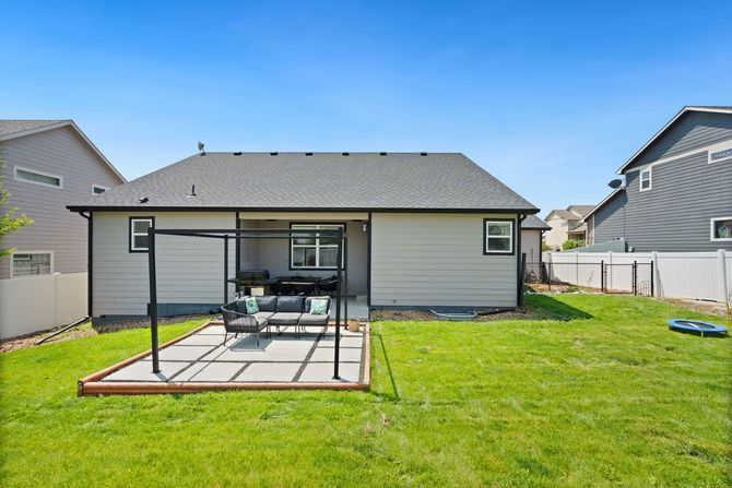 Backyard patio with pergola, outdoor table, and chairs beside a house on a grassy lawn.