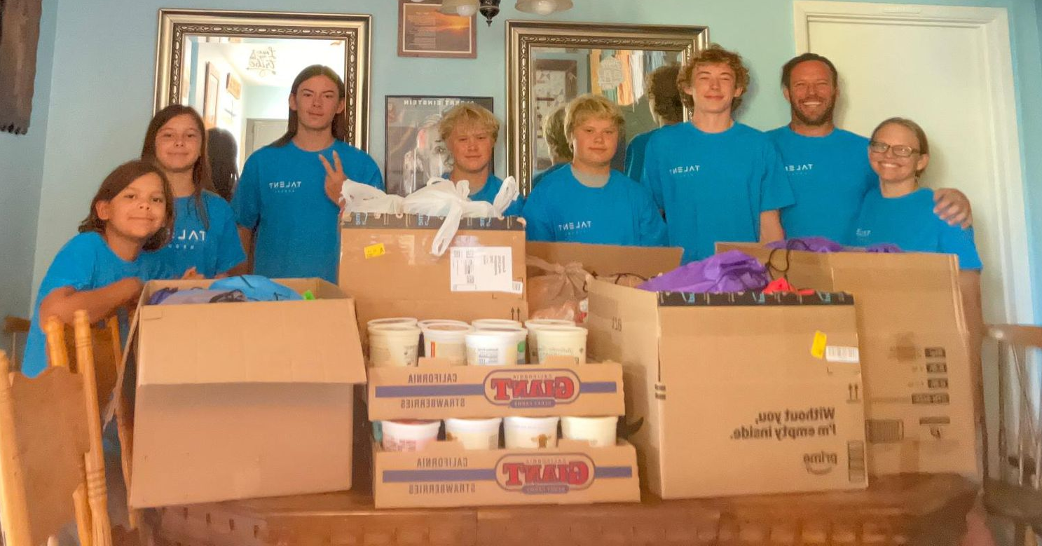 Group of volunteers in blue shirts standing behind boxes of donated food in a home interior