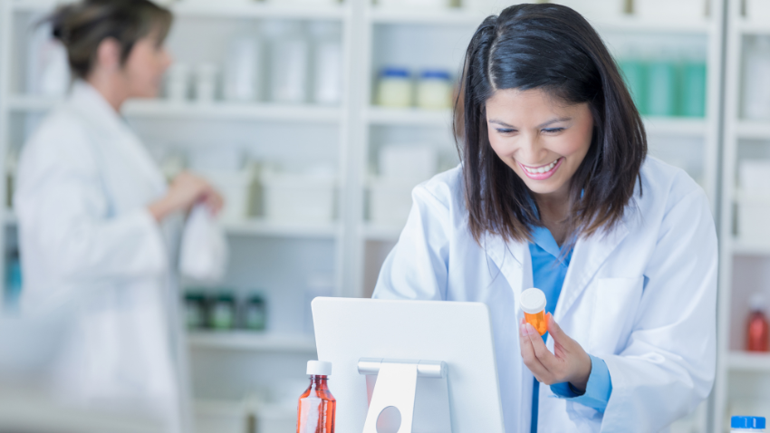 A pharmacist in a lab coat checks a medication bottle while working at a computer in a pharmacy.