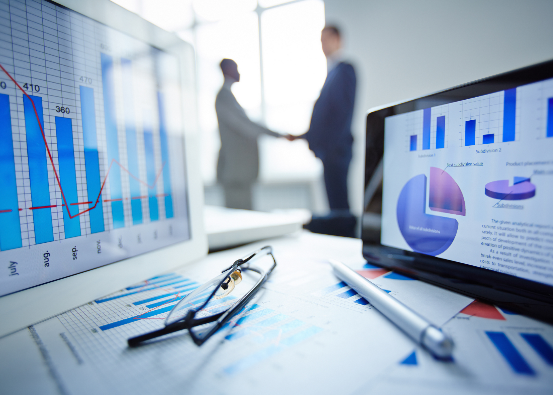 Business meeting: charts and graphs on tablets and papers, two people shaking hands in background.