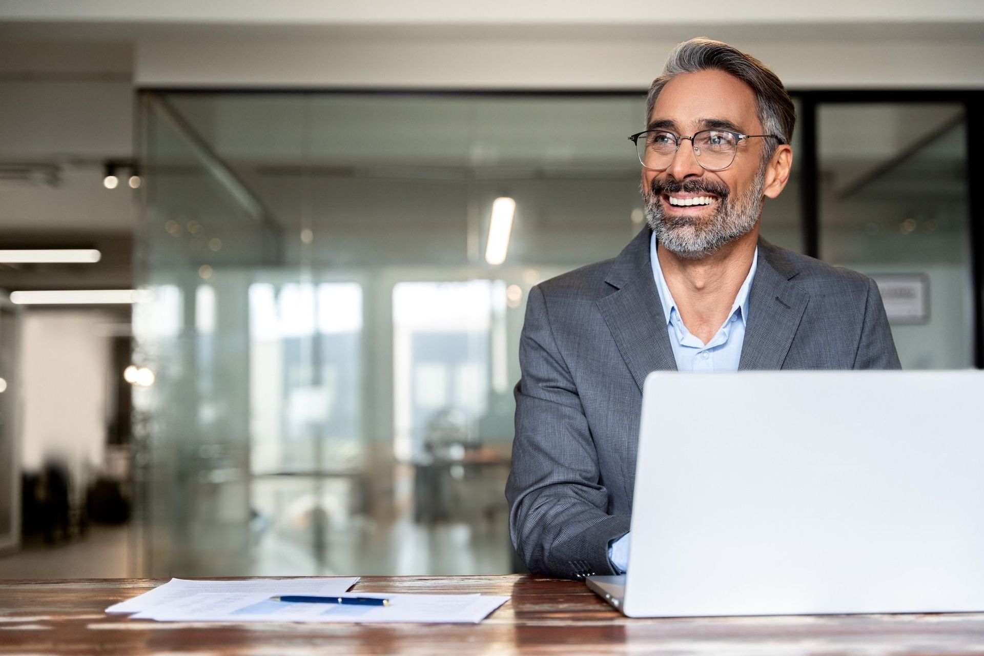 A smiling person in a grey blazer and glasses works on a laptop at a wooden desk in a modern office.