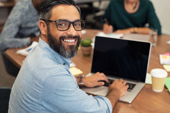 A smiling man in a blue shirt works on a laptop at a table during a meeting.