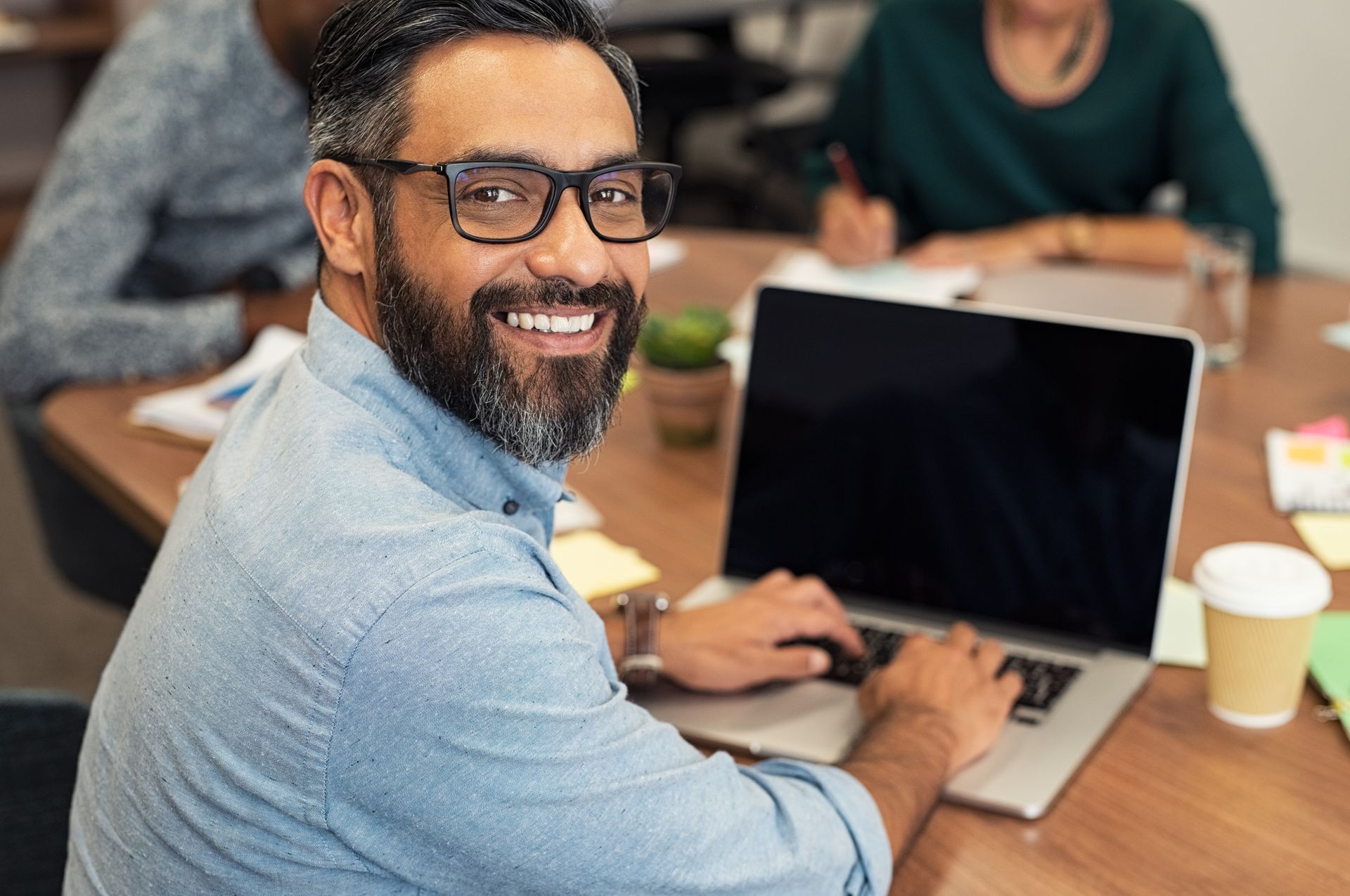 Man with glasses smiles at the camera while typing on a laptop at a table, people in background.