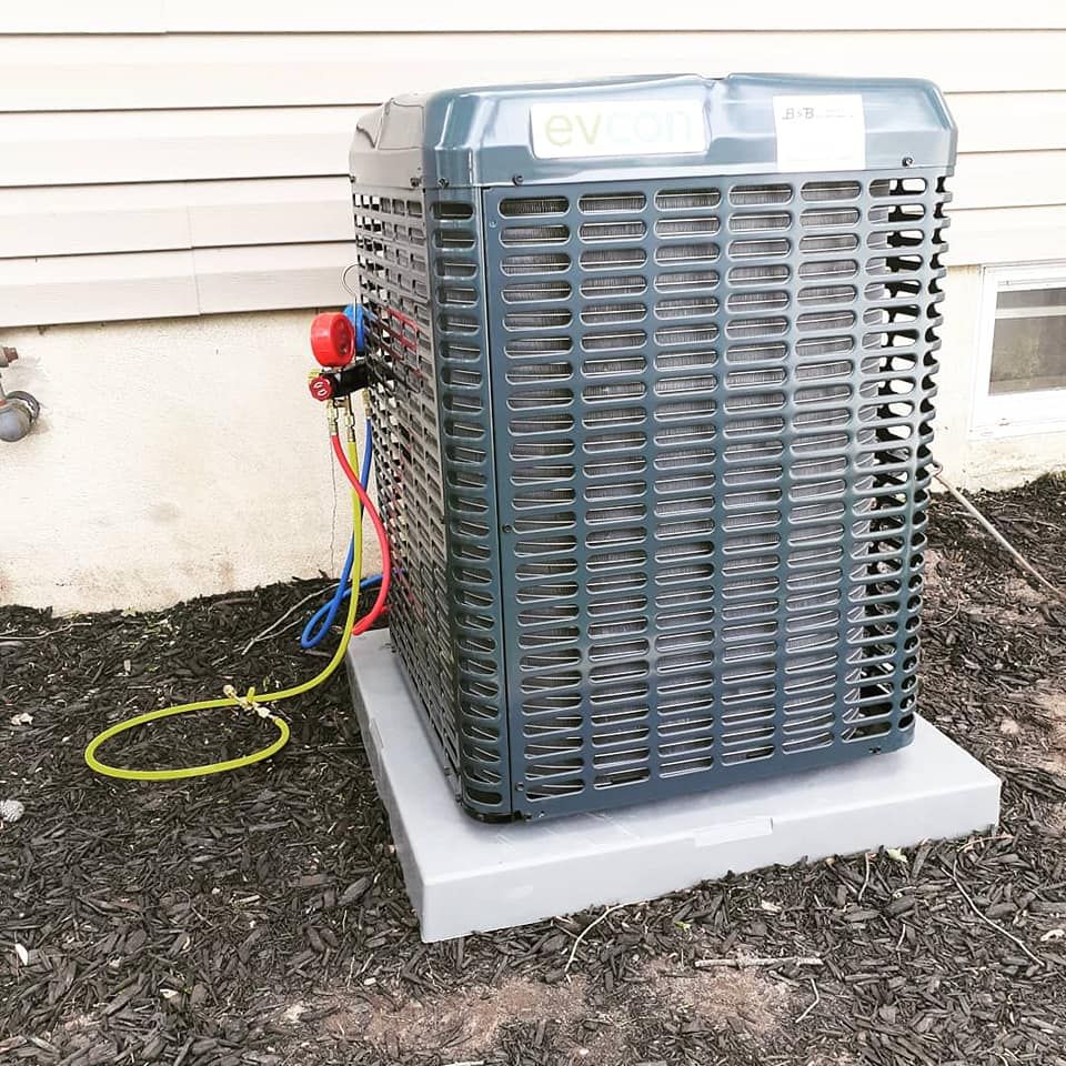 AC unit outside on a gray base, with gauges attached. Set in a yard with dark mulch.