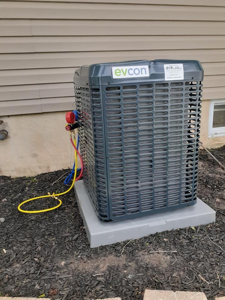 An Evcon air conditioning unit sits on a gray pad on a bed of dark mulch. Red and blue hoses connect to it.