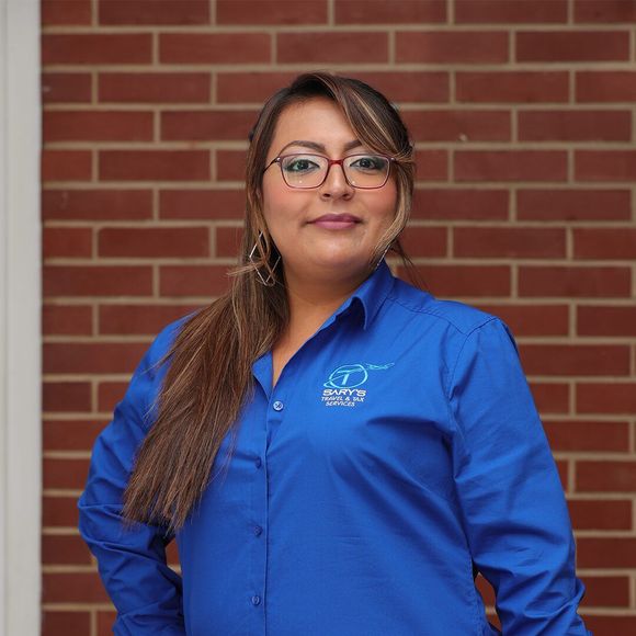 Woman in blue button-up shirt standing against a brick wall, wearing glasses and smiling.