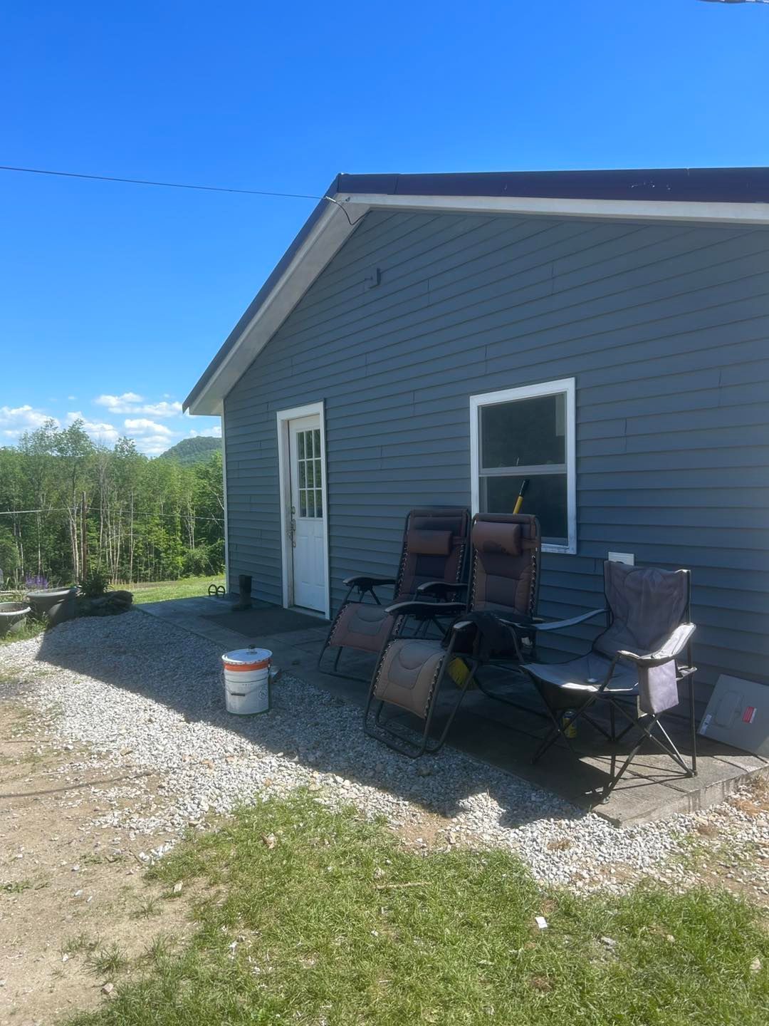 Gray house exterior with white-trimmed door and window, gravel yard, and stacked patio chairs by the wall