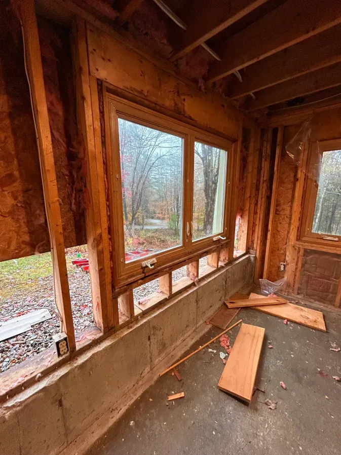 Interior of unfinished wood-framed room with large windows, exposed studs, and loose boards on the floor