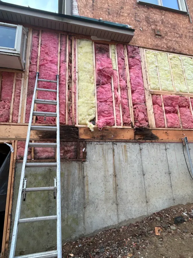 House exterior under renovation with pink and yellow insulation exposed and a ladder leaning against the wall