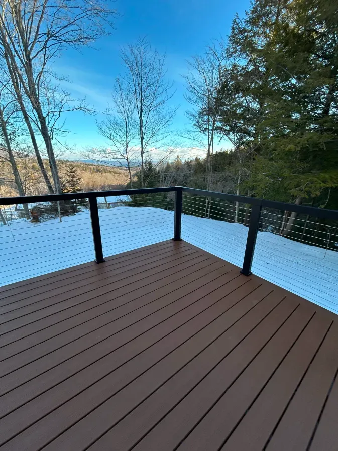 Snow-covered wooden deck with black railing overlooking a wooded landscape under a blue sky
