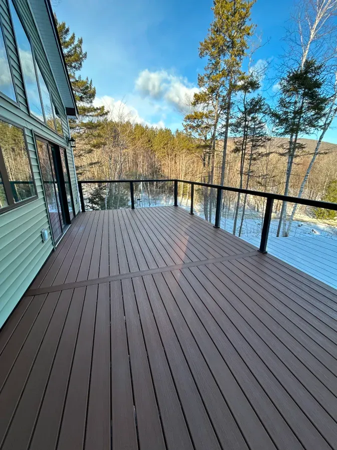 Spacious wooden deck beside a house, overlooking snowy ground and trees under a blue sky.