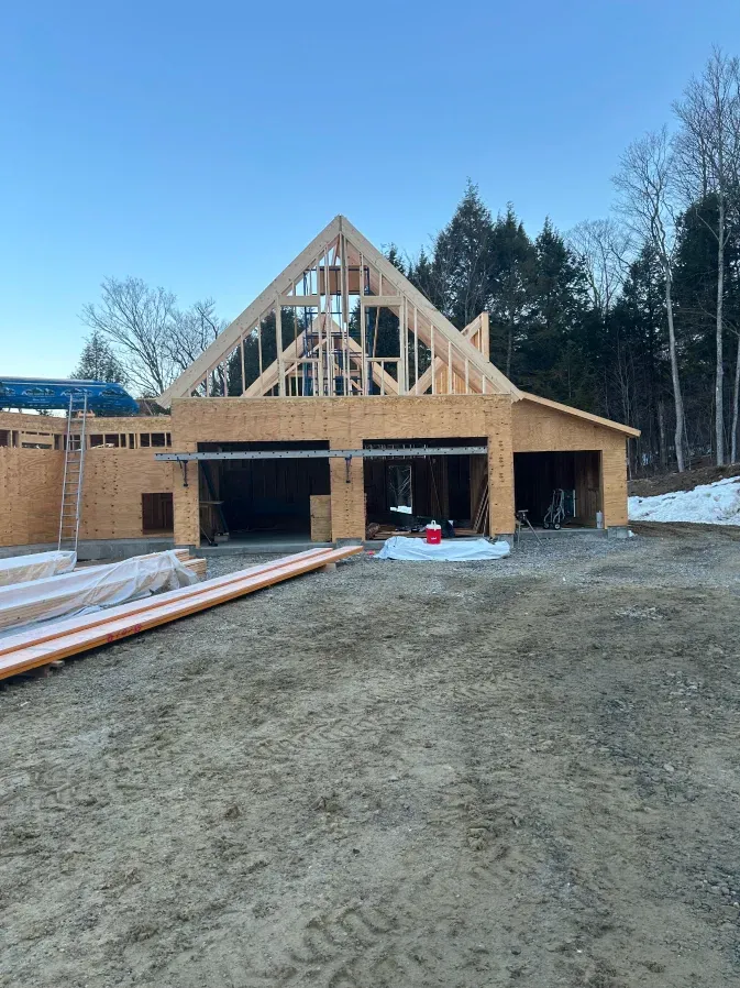 Wood-framed house under construction with exposed roof trusses and open garage on a gravel lot.