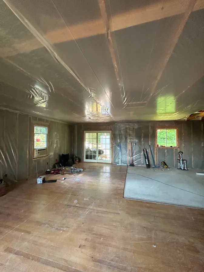 Large unfinished room with exposed insulation, wood floors, and tools near a window and glass doors.
