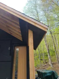 Cabin corner with wood beam and slanted roof beside a forest, with a tarp-covered object below