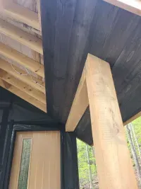 Wooden porch roof and support beam beside a door, with exposed rafters and green trees outside.