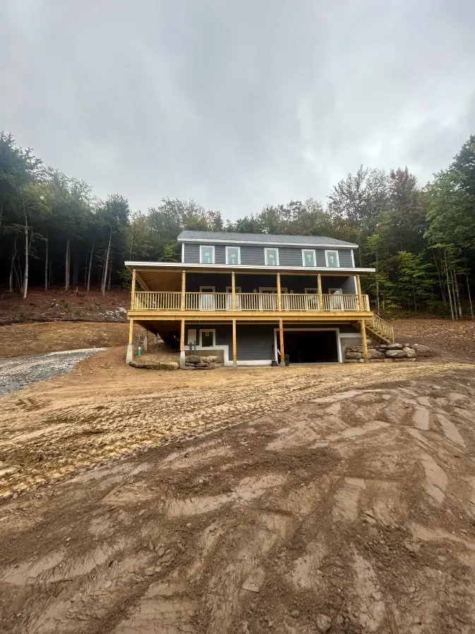 New house on a muddy clearing, surrounded by trees, with a two-story porch and gray roof
