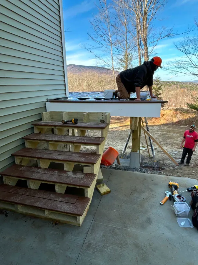 Person installing a railing on a wooden deck beside house steps, with child nearby and tools on concrete patio