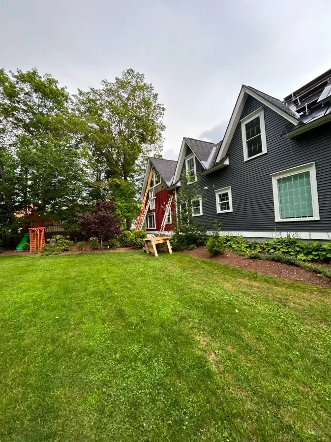 Green lawn beside a dark gray house with white trim, trees, and a red porch in the background