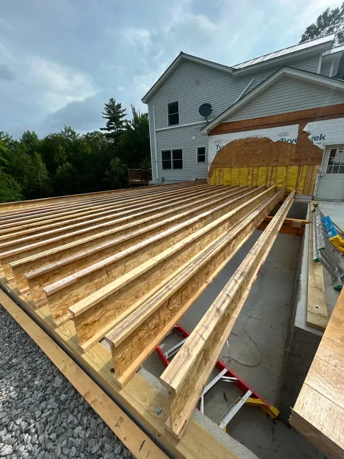 Wooden floor joists installed on a house addition beside a white two-story home under a cloudy sky