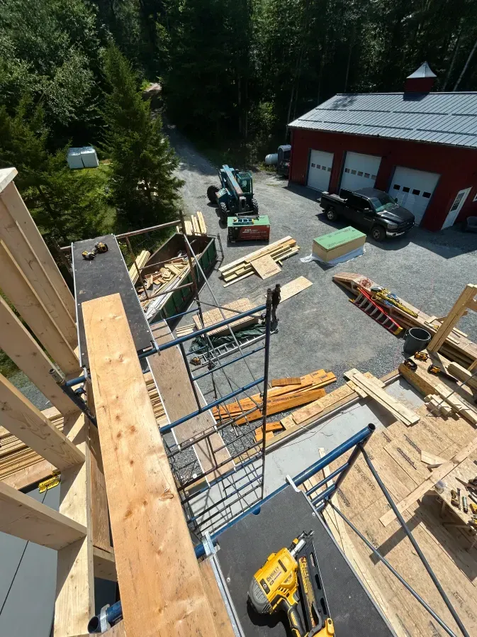 Roof construction site with ladders, scaffolding, lumber, and a red garage beside trees.
