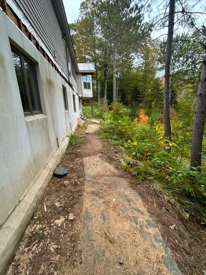 Narrow dirt path beside a white house leading through trees and greenery in a wooded yard