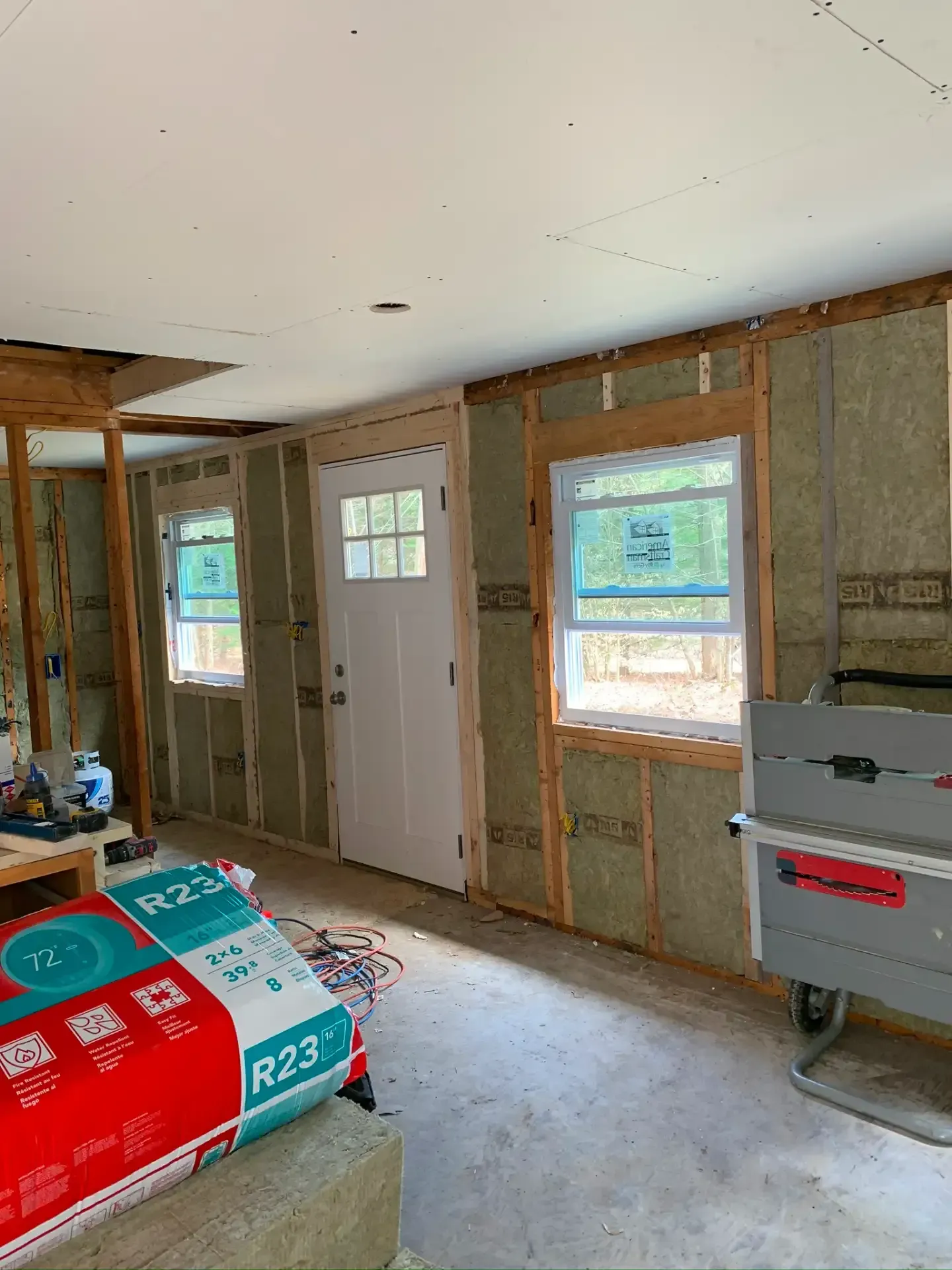 Unfinished interior room with insulation, exposed framing, windows, a white door, and construction materials.