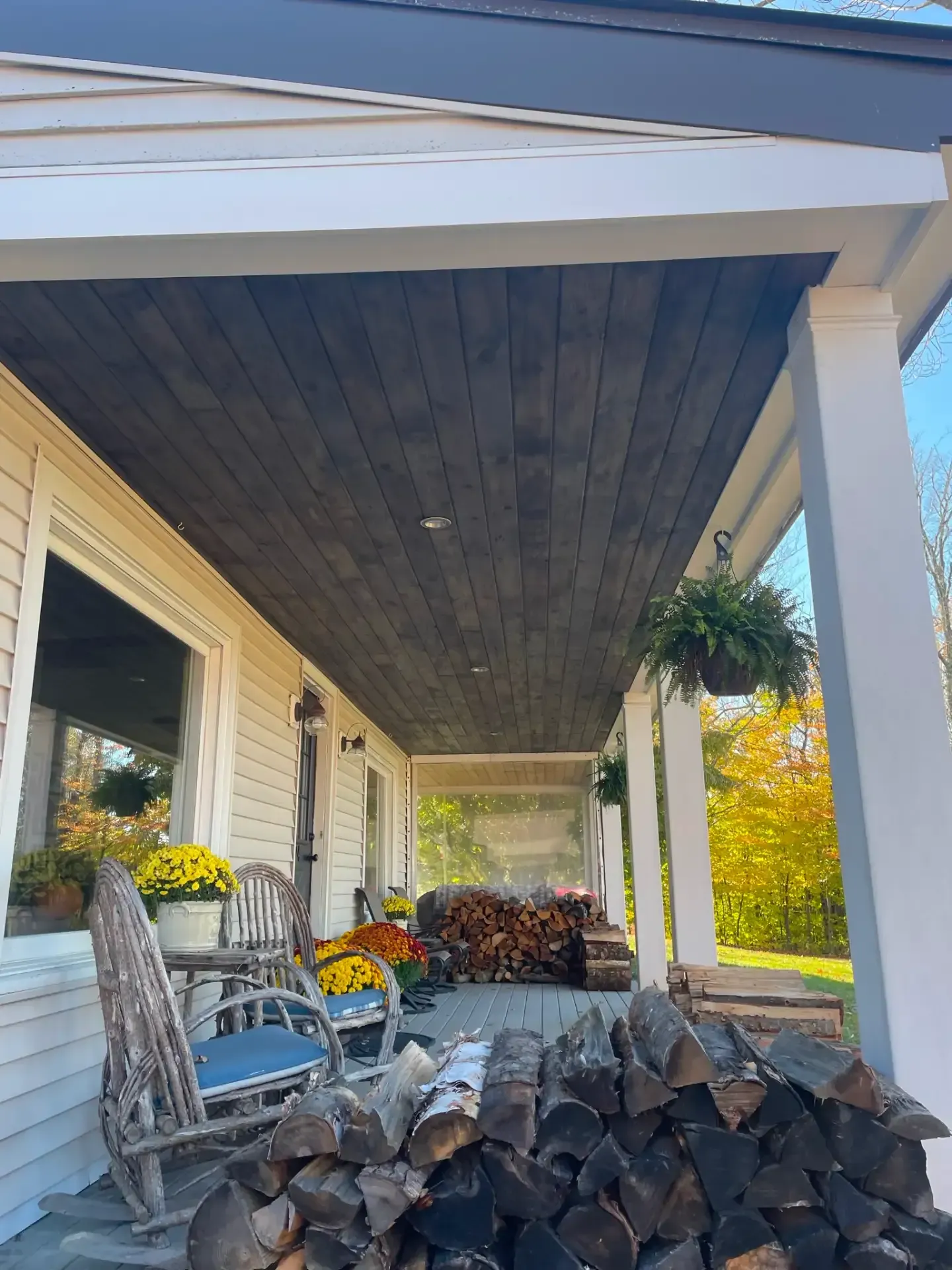 Porch with stacked firewood, rocking chairs, hanging plant, and view of yard and trees