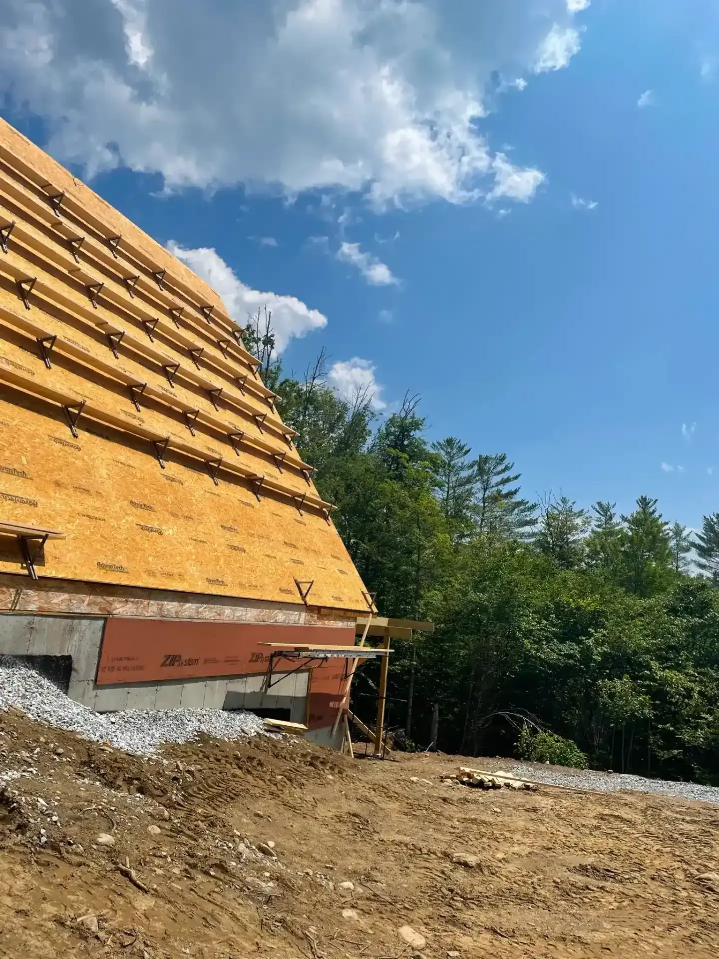Partially built house with exposed wooden roof framing beside a dirt lot and trees under a blue sky.