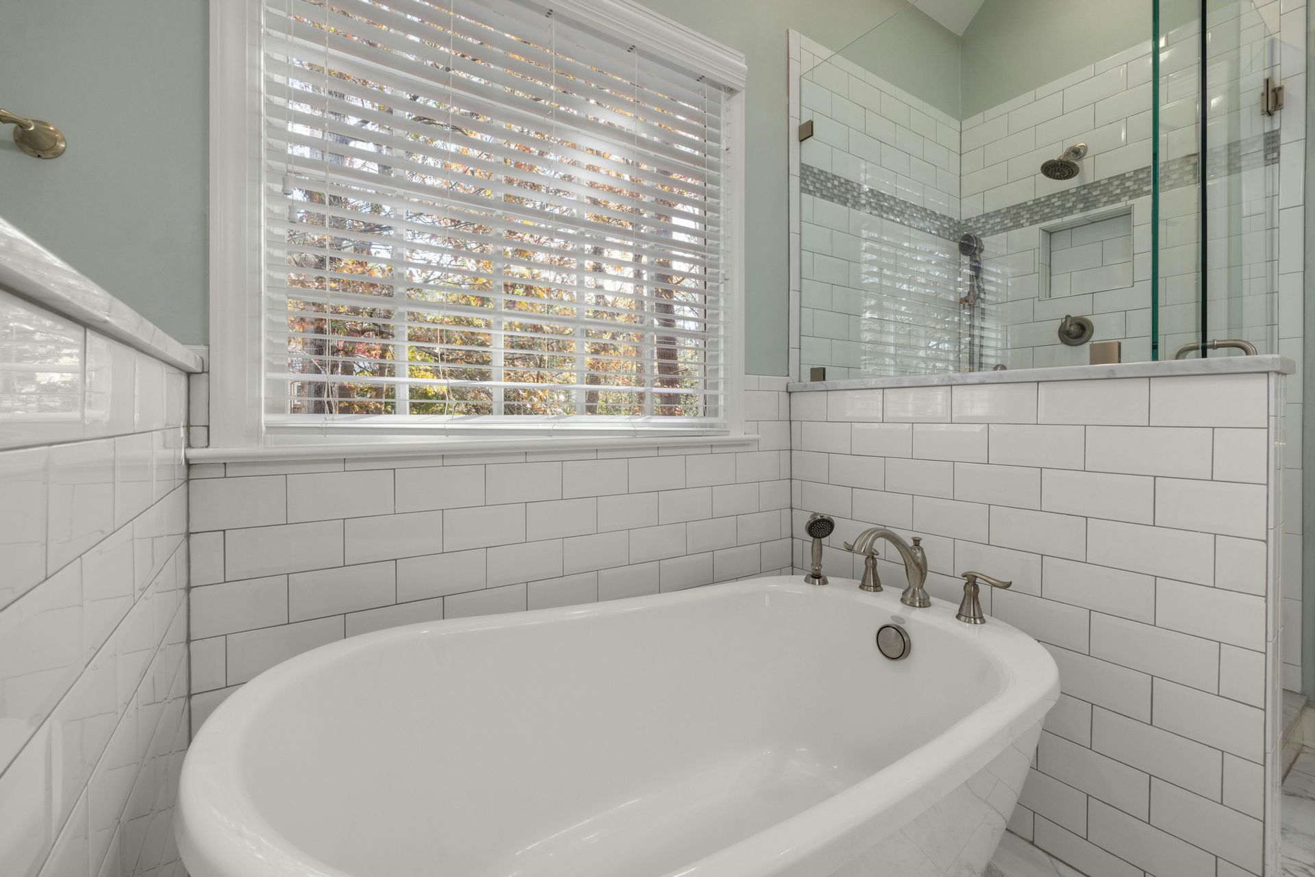 Bright white bathroom with a freestanding tub, tiled walls, window blinds, and glass shower enclosure