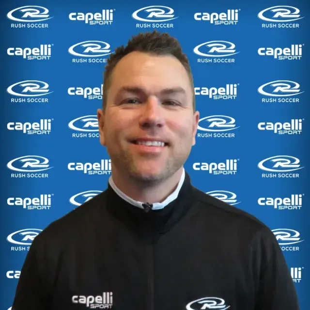 Man in black Capelli top smiling in front of a blue Rush Soccer sponsor backdrop