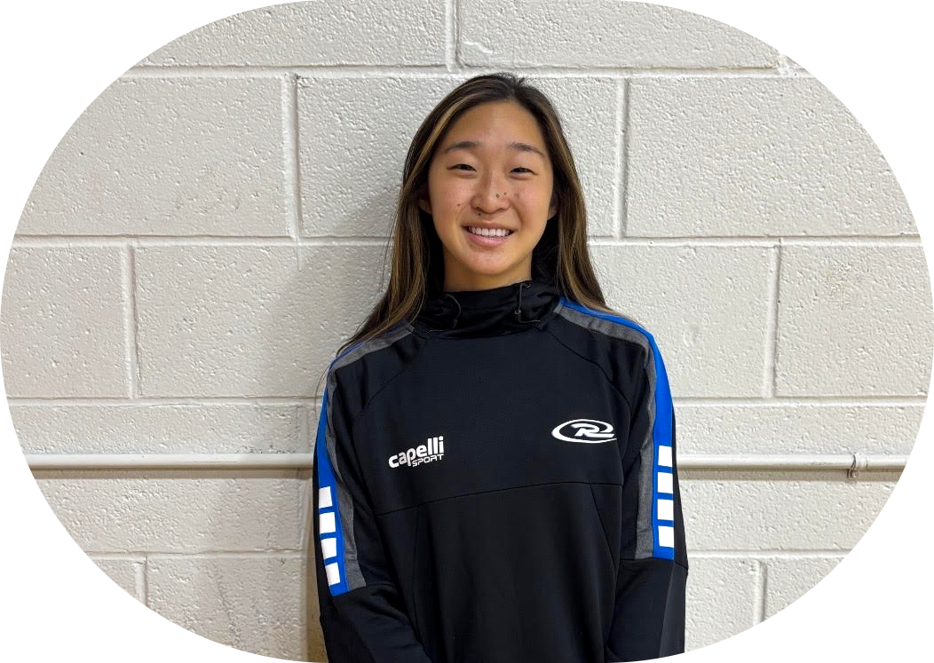 Person smiling in a black athletic jacket with blue sleeves, standing against a light gray brick wall.