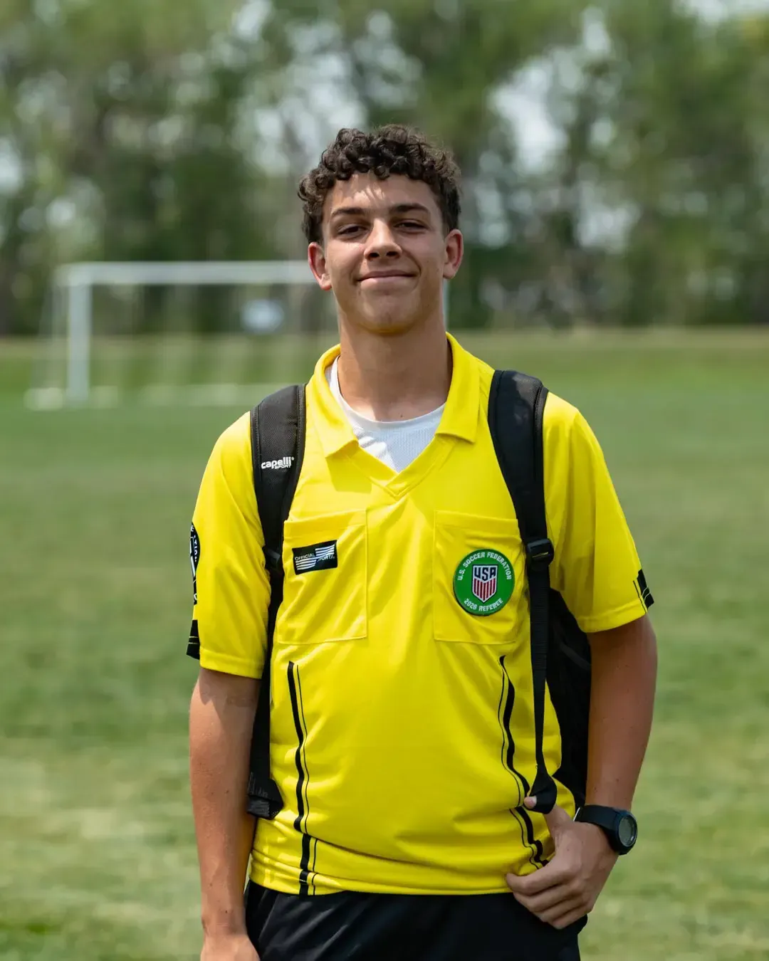 Person in a bright yellow polo shirt and backpack standing on a soccer field with a goal in the background.