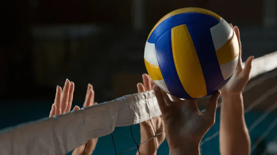 Volleyball hitting at the net, blue and yellow ball above players' hands in a gym
